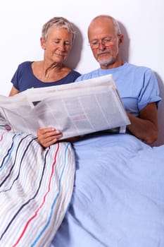 Senior couple with husband and wife lie in bed in the morning evening together with a tablet and a newspaper