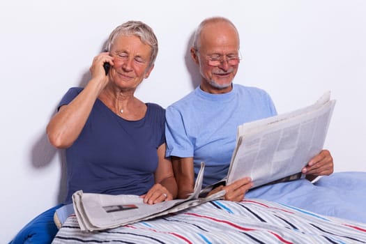 Senior couple with husband and wife lie in bed in the morning evening together with a tablet and a newspaper
