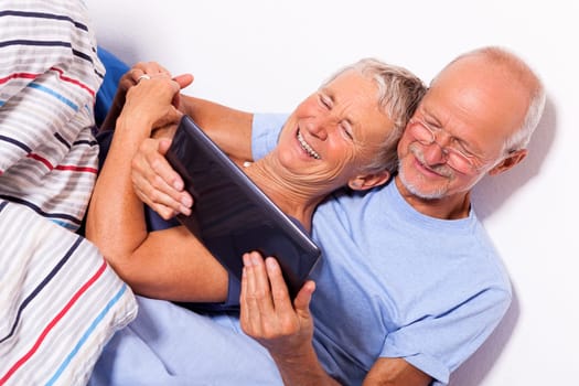 Senior couple with husband and wife lie in bed in the morning evening together with a tablet and a newspaper