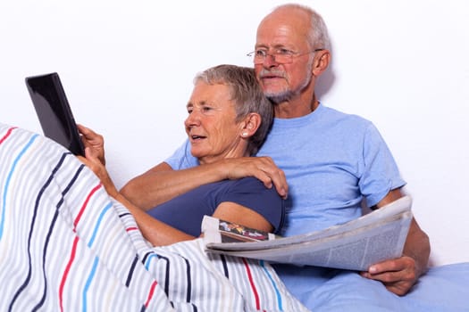 Senior couple with husband and wife lie in bed in the morning evening together with a tablet and a newspaper