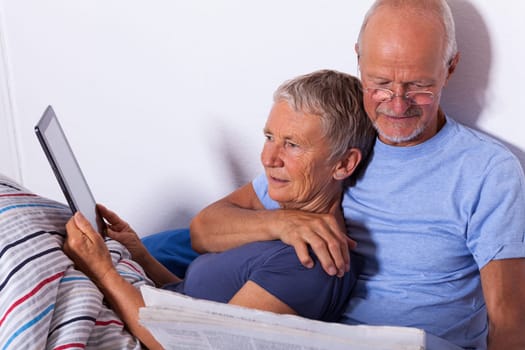 Senior couple with husband and wife lie in bed in the morning evening together with a tablet and a newspaper