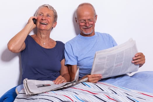 Senior couple with husband and wife lie in bed in the morning evening together with a tablet and a newspaper