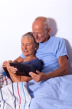 Senior couple with husband and wife lie in bed in the morning evening together with a tablet and a newspaper