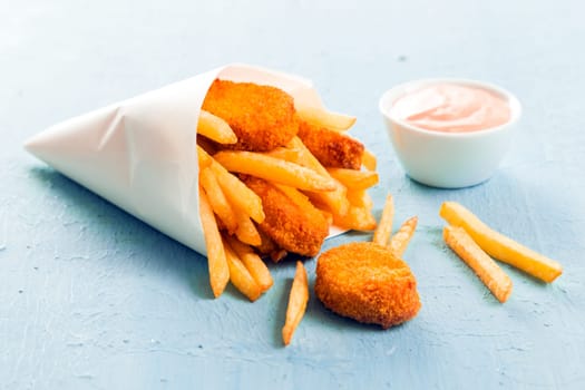 Golden fish nuggets with fried potato chips or French fries spilling out of a takeaway paper cone onto a blue surface with a small bowl of mayo dip