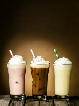 Three refreshing creamy milkshakes in berry, chocolate and lemon flavors topped with a twirl of ice cream or frozen yoghurt on a wooden table against a brown background with copyspace