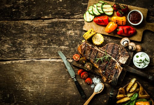 Overhead view of colorful roast vegetables, savory sauces and salt served with grilled t-bone steak on a rustic wooden counter in a country steakhouse
