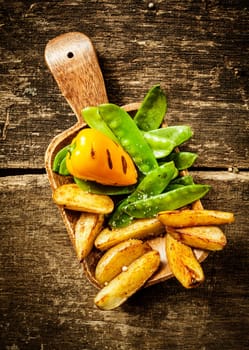 Side serving of delicious cooked vegetables served on a wooden spatula with roasted sweet red pepper, green mangetout peas and potato wedges on a rustic grunge wooden background