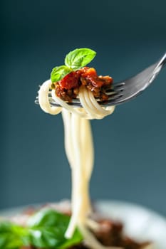 Fork full of twirled Italian spaghetti with a Bolognese meat sauce and basil suspended in the air above a full plate of food against a grey background