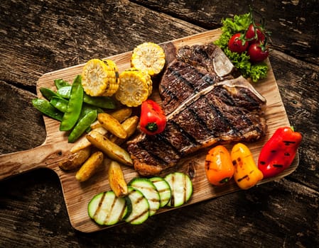 Delicious steakhouse porterhouse steak and colorful fresh roast vegetables with mangetout peas, corn, zucchini, bell pepper, potato and tomato on a wooden board, view from above