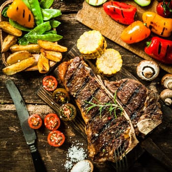 View from above of a delicious grilled porterhouse steak with assorted roast vegetables including tomato, peppers, mushrooms, corn, mangetout, and potato wedges