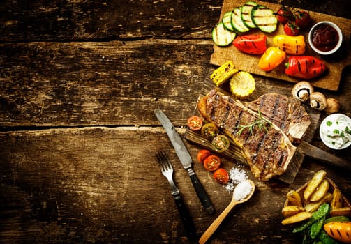 Preparing t-bone steak and roast vegetables in a country kitchen with bell peppers, mushrooms, tomato, potato, mangetout peas, and corn on a rustic wooden table with copyspace