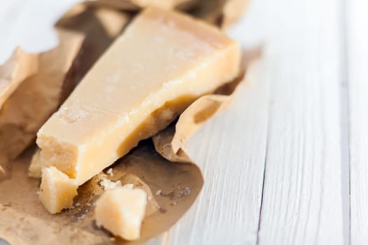 Wedge of hardy crumbly matured Italian Parmigiano-Reggiano cheese, or Parmesan, in a torn brown paper wrapper on a white wooden table
