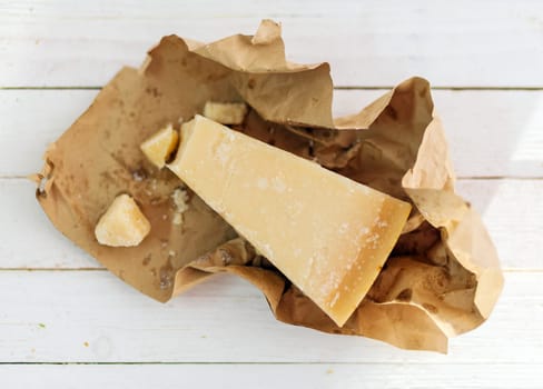 Wedge of hard granular Parmigiano-Reggiano cheese used extensively in Italian cuisine in a brown paper wrapper on white boards viewed from overhead
