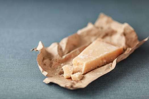 Wedge of hard Italian Parmigian-reggiano cheese on a torn brown paper wrapper for use as a cooking ingredient on a grey textile with copyspace