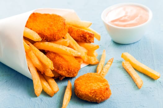 Fried fish nuggets with golden French fries spilling out of a paper cone from a takeaway fish shop onto a blue surface, close up view