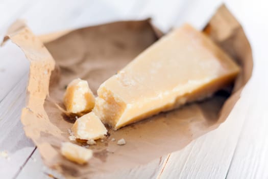 Wedge of fresh matured Italian Parmesan cheese or Parmigiano-Reggiano in a torn brown paper wrapper on white boards for use in Mediterranean cuisine