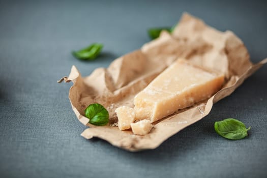 Portion of mature Italian Parmesan cheese with a traditional hard granular texture with scattered fresh basil on crinmpled brown paper, on a grey cloth with copyspace