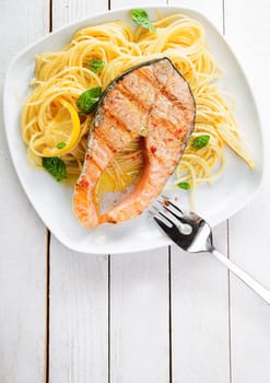 Gourmet seafood cuisine with grilled salmon cutlet steak served on a bed of Italian linguine pasta garnished with lemon and fresh basil, overhead view on a white wooden table with copyspace