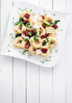 Tortellini pasta with fresh herbs, a spicy piquant sauce and feta cheese on an elegant plate on a white wooden table with copyspace