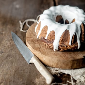 Freshly baked glazed ring cake with lemon icing on a round wooden board with a large kitchen knife to serve it in square format in a rustic setting