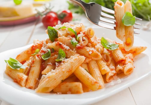 Delicious savory Italian penne rigate pasta with fresh basil and grated parmesan cheese served with a salad with a fork with two pasta tubes suspended above the plate