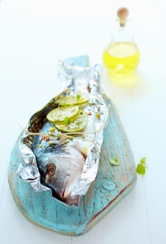 Preparing a whole fresh fish to be baked on a barbecue in a foil wrapper with lemon, olive oil , spices and herbs arranged on a wooden board with the head to the camera