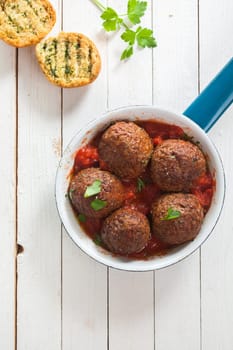 Tasty savory ground beef meatballs in tomato sauce served in a saucepan with crusty toasted herb bread on a white rustic wooden table, overhead view