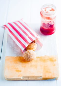 Colorful striped red and white paper takeaway packet of fresh sugared mini doughnuts on a wooden bread board with a bottle of jam or preserve behind