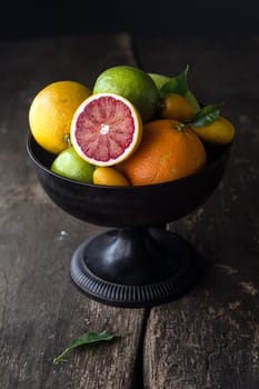 Pedestal bowl of assorted fresh citrus fruit with a halved blood orange displayed at the front on a rustic wooden background with shadows