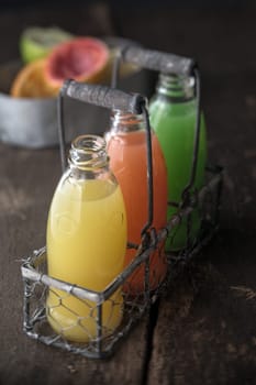 Three full bottles of natural juice, yellow, orange and green, made from squeezed citrus fruits as lemon, grapefruit and green pomelo, in a metallic rustic basket, on a wooden table