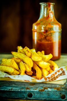 Salty French fried potatoes on a paper next to a bottle of homemade ketchup or tomato sauce, on a wooden old rustic table