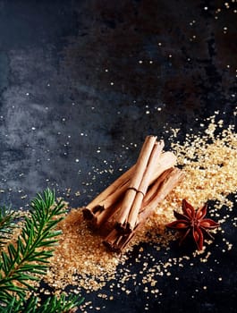 Christmas background with traditional spices with a bundle of stick cinnamon and star anise on scattered caramelized brown sugar scattered on a dark surface with copyspace and decorated with pine