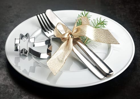 Festive Christmas place setting with a shiny silver shooting star cookie cutter alongside a knife and fork tied with a decorative gold ribbon with a sprig of pine foliage on a plain white plate