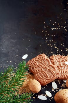 Traditional crunchy spicy speculoos Dutch Christmas buscuits originally baked for the feast of St Nicholas with flaked and whole almonds and a pine branch, on a dark backgorund with copyspace