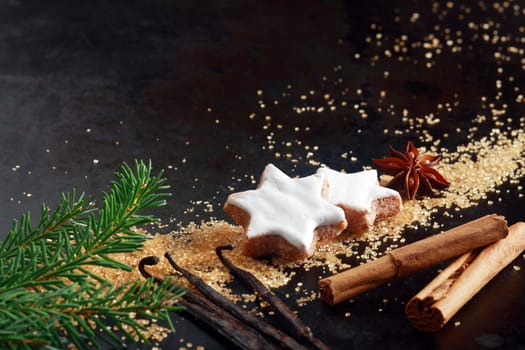 Crunchy spicy star shaped Christmas biscuits glazed with white icing arranged on a dark background with sugar granules, star anise, cinnamon and vanilla pods, with copyspace