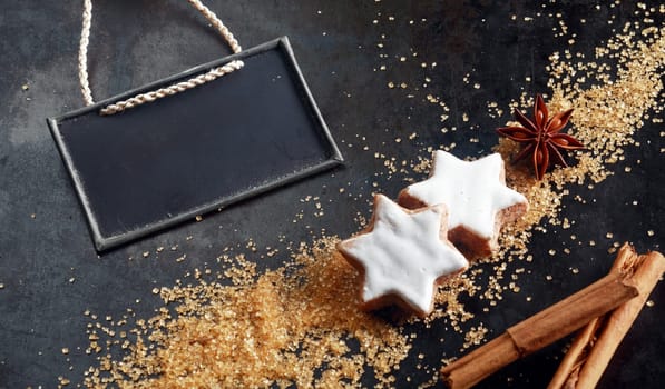 Christmas baking background with glazed white spicy star shaped biscuits surrounded by ingredients - star anise, brown sugar and stick cinnamon with a blank slate with copyspace for a menu or recipe