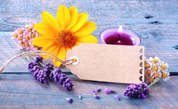 Fresh floral wellness and spa still life with spikes of scented lavender, dainty blosoms and a colourful yellow gerbera surrounding a blank gift tag with copyspace and a burning aromatherapy candle