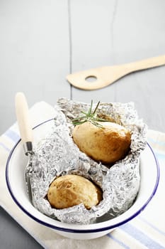 Fresh hot baked potatoes with rosemary still nestling in their foil wrappers in a metal basin on a white painted wooden picnic table, with copyspace