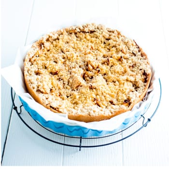 Delicious baked plum pie with a golden pastry crumble topping standing on a wire rack ready to be served on white wooden boards, square format