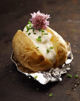Barbecued baked potato on its foil wrapper served with a filling of sour cream, chopped fresh green chives and a chive flower on a dark background