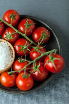 Bunch of ripe red cherry tomatoes on the vine arranged on a plate with a bowl of coarse natural sea salt for use as cooking ingredients in the kitchen