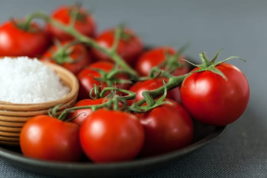 Close up low angle view of a plate of fresh ripe red cherry tomatoes on the vine with a small bowl of coarse sea salt ready to be used as ingredients in cooking