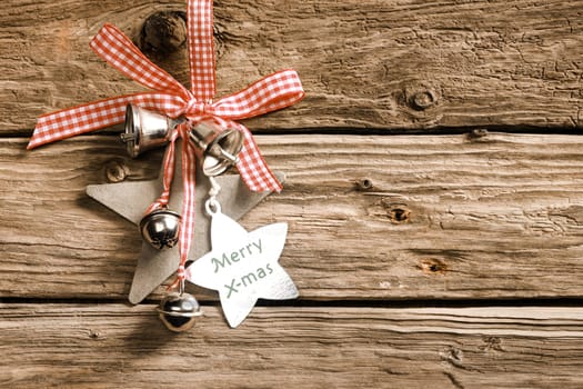 Celebrating a country Christmas with decorative stars and silver bells habging from a red and white checked ribbon and bow on rustic wooden boards with a star-shaped tag with the message - Merry X-mas
