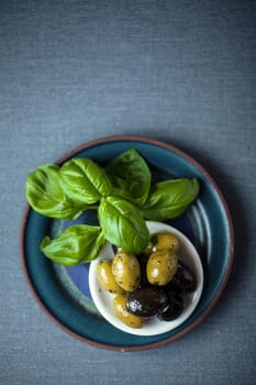 Overhead view of a bunch of fresh basil leaves with a bowl of black and green cured olives on a grey weave textile background with copyspace