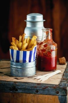 Colorful blue striped packet of crispy of French fries standing on a rustic wooden table in a fish and chip shop with condiments