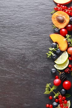 Border of fresh fruit and herbs along the right of the frame with assorted berries, apricot, peach and lemon on a dark grey slate background with copyspace