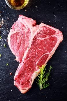 Uncooked t-bone or porterhouse steak seasoned with salt and spices with fresh herbs with a glass of whiskey above