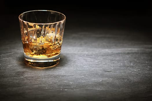 Whiskey on the rocks in a glass tumbler standing on a bar counter in the shadows with a highlight over copyspace or a place for product placement