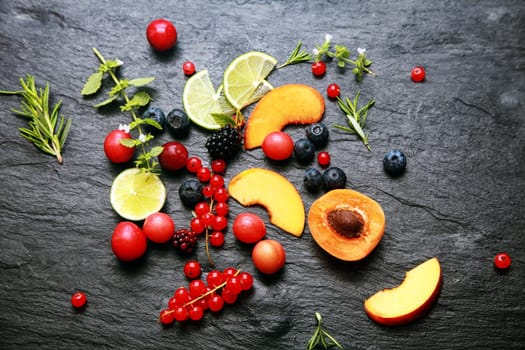 Scattered fresh fruit and berries with cranberries, blackberries, redcurrants, lemon, peach, blueberries, rosemary and herbs on a textured slate background