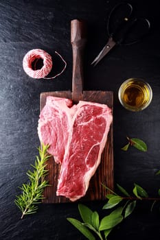 Seasoning a t-bone steak for cooking with bay leaves, fresh rosemary, olive oil and string, overhead view on a wooden board on a rustic kitchen counter
